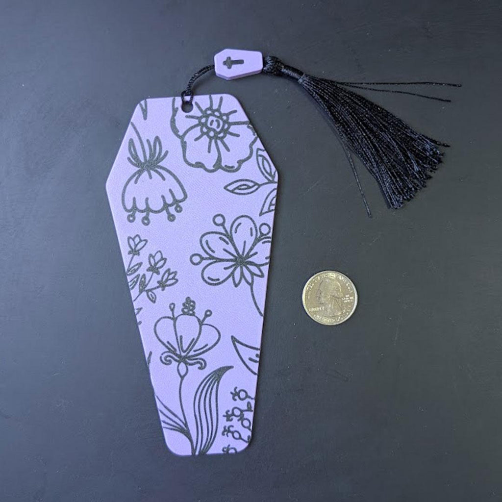 Decorative bookmark with floral patterns and a tassel on a dark surface, next to a coin for scale.