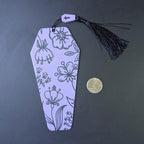 Decorative bookmark with floral patterns and a tassel on a dark surface, next to a coin for scale.