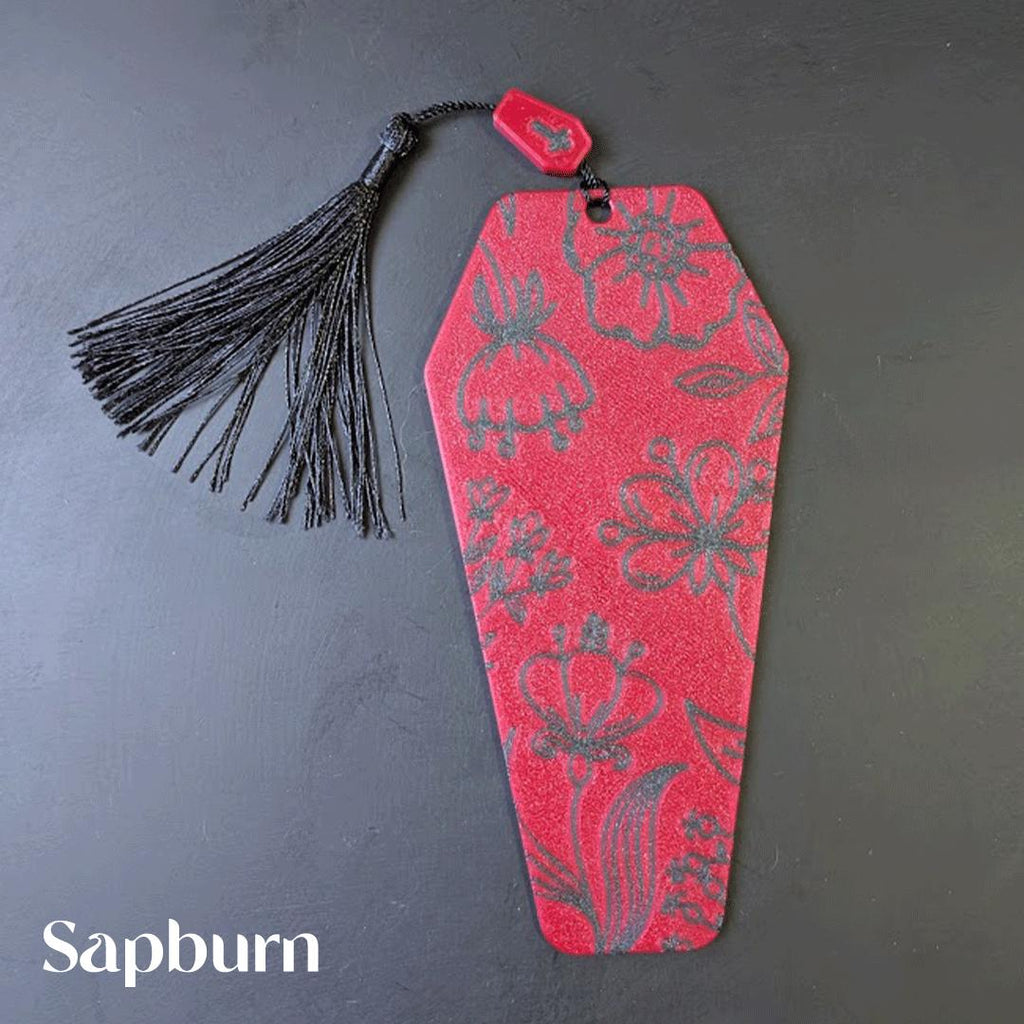 Red coffin-shaped bookmark with black floral pattern and tassel on a dark background, featuring the brand 'Sapburn'.