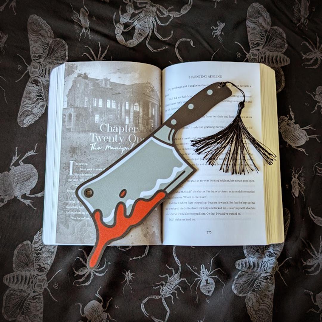Open book with a bookmark of a cleaver and tassel on a dark fabric background with insect illustrations.